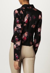 Femme portant une veste noire ajustée à imprimé floral multicolore et un pantalon beige, vue de dos sur un fond uni.