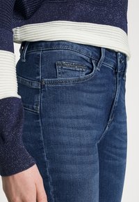 Primo piano di una persona che indossa jeans blu scuro con cucitura decorativa sulla tasca e un maglione a maglia blu navy e bianco glitterato.
