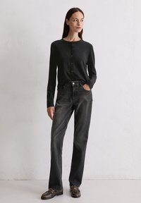 Cardigan noir à boutons avec des manches longues, associé à un jean droit gris foncé. Le modèle porte des chaussures à boucle marron foncé. Design simple.