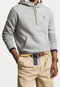 Persona che indossa un felpa grigia chiara con tasca frontale, pantaloni beige con cintura a righe, una maglietta gialla sotto e un orologio al polso sinistro.