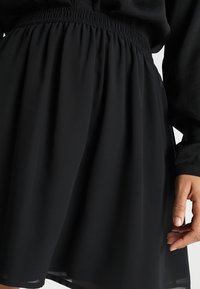 Černá sukně s elastickým pasem nošená s černým topem s dlouhým rukávem, ruka uvolněně podél boku, látka shromážděná v pase.