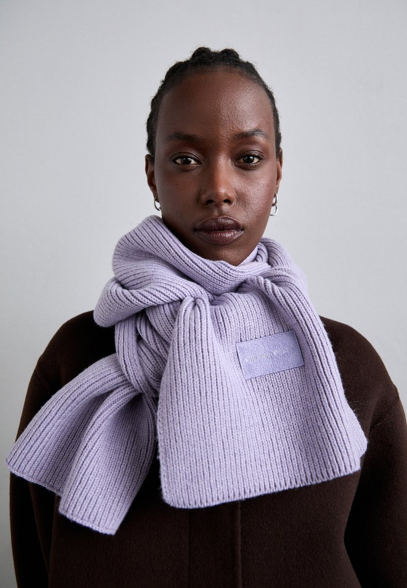 Lawendowy szal z ribbedu, luźno zarzucony, z prostokątną metką z wytłoczonym tekstem, noszony na ciemno-brązowym płaszczu.