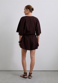 Mujer con el cabello recogido en un moño, vistiendo un vestido suelto marrón oscuro y sandalias con tiras tobilleras adornadas, de pie de espaldas a la cámara.