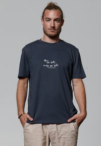Navyblauwe katoenen T-shirt met korte mouwen, met witte tekst op de voorkant. De tekst zegt: "Es ist, wie es ist." Het model draagt lichte broek.