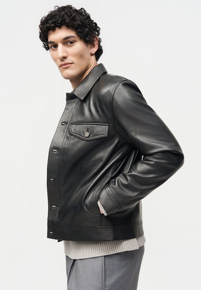 Άνδρας με σγουρά σκούρα μαλλιά, που φοράει μαύρο δερμάτινο μπουφάν, ανοιχτό γκρι πουλόβερ και γκρι παντελόνι σε απλό λευκό φόντο.