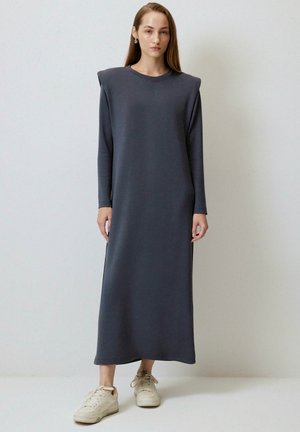 Kobieta stoi przy gładkiej ścianie, ubrana w długą, szarą sukienkę maxi z poduszkami na ramionach oraz kremowe trampki, patrzy przed siebie.