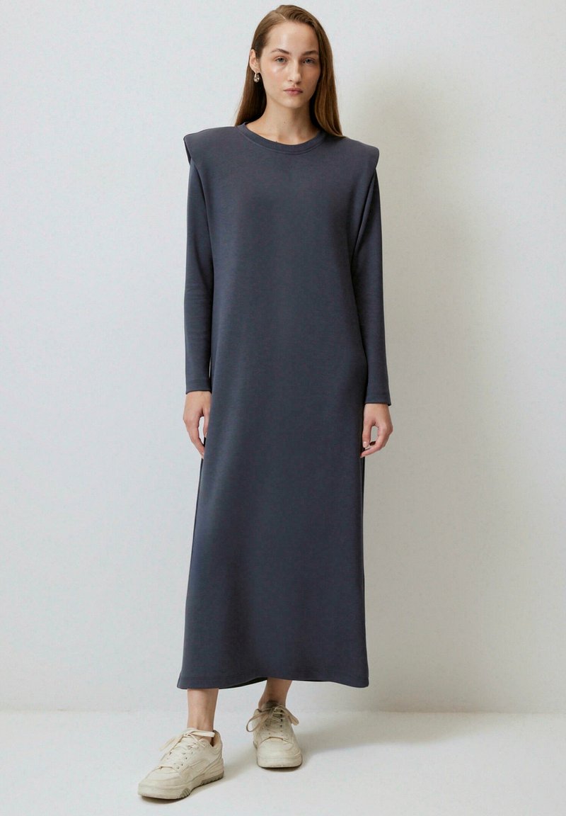 Femme debout contre un mur uni portant une robe longue gris foncé à manches longues avec épaulettes et des baskets blanc cassé, regardant droit devant.