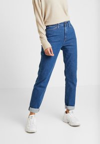 Jean en denim bleu avec une coupe droite, présentant des ourlets roulés et un design classique à cinq poches. Associé à un pull en texture beige et des baskets blanches.
