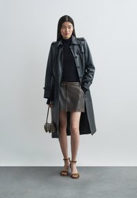 Abrigo de trench negro de cuero sobre una blusa oscura, combinado con una falda corta de cuero marrón. Los accesorios incluyen un bolso pequeño y sandalias de tacón verdes.