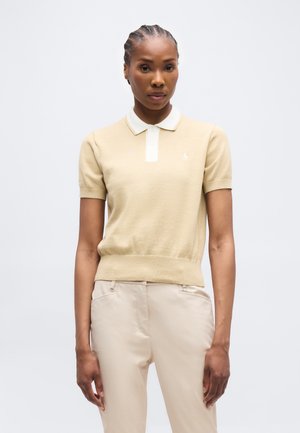 Γυναίκα με πλεξούδες, ντυμένη με μπεζ πόλο μπλουζάκι με κοντά μανίκια και ανοιχτό μπεζ παντελόνι, στέκεται μπροστά σε απλό λευκό φόντο.