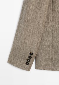 Brown tweed jacket sleeve with a textured finish, featuring four black buttons and subtle stitching detail at the cuff.