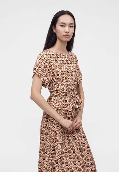 Femme aux longs cheveux noirs portant une robe à manches courtes avec un motif floral rose, marron et noir, et une ceinture nouée à la taille, debout les mains jointes.
