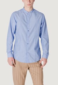 Camicia a maniche lunghe azzurra con colletto alla cinese, dotata di chiusura frontale con bottoni e una sottile texture. Abbinata a pantaloni beige a righe.