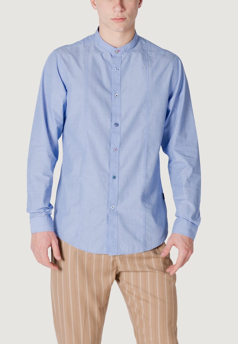 Camicia a maniche lunghe azzurra con colletto alla cinese, dotata di chiusura frontale con bottoni e una sottile texture. Abbinata a pantaloni beige a righe.