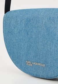 Borsa a tracolla in denim blu con forma ovale, dotata di una tracolla nera e un accento argentato con logo vicino alla parte inferiore. Texture liscia.