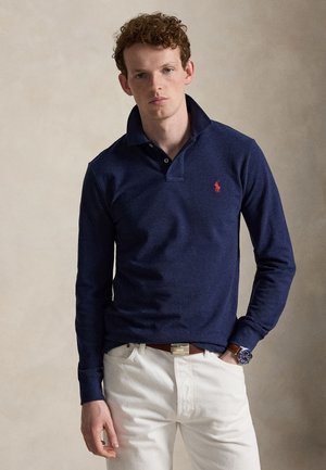 Jeune homme aux cheveux bouclés portant un polo bleu marine à manches longues et un pantalon blanc, debout avec une main dans la poche devant un fond neutre.