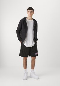 Sudadera negra con cremallera sobre una camiseta blanca, combinada con pantalones cortos de malla negros con un parche de logo, zapatillas deportivas blancas de caña alta y calcetines.