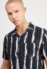 Camisa negra de manga corta con patrón de rayas blancas, botones en la parte delantera y un solo bolsillo en el pecho. Hecha de una tela ligera.