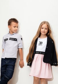 Due bambini posano davanti a un muro bianco. Il ragazzo indossa una maglietta grigia con una banda di logo nera e jeans scuri. La ragazza ha i capelli lunghi, una maglietta bianca e una gonna rosa con una banda di logo nera.