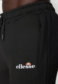 Schwarze Sporthose mit elastischem Bündchen, Kordelzug, Reißverschlusstaschen und dem weißen "ellesse"-Logo mit farbenfrohen Akzenten.