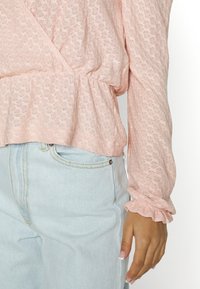 Blusa texturizada rosa claro com cintura franzida e punhos de mangas com babados, combinada com jeans de cintura alta azul claro com costura amarela.