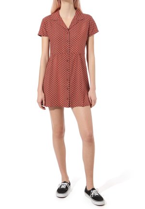 Frau trägt ein rotes kurzärmeliges Kleid mit weißen Blumenmustern und schwarze Schnür-Sneaker, steht vor weißem Hintergrund.