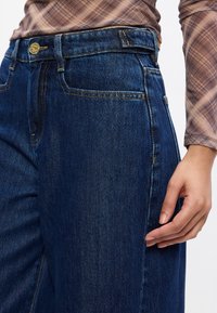 Jeans de ganga azul de cintura alta, apresentando um design de perna reta, costura contrastante e discretos detalhes em metal na parte da frente.