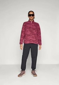 Φλις πουλόβερ σε σκούρο ροζ με σχέδιο ζιγκ-ζαγκ, με μισό φερμουάρ στον γιακά; συνδυασμένο με μαύρο παντελόνι και καφέ πεζοπορικά παπούτσια. Το μοντέλο φορά σκούρα γυαλιά ηλίου.