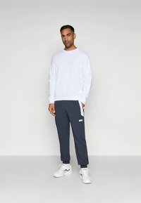 Hombre de pie frente a un fondo liso, vistiendo un sudadera blanca, pantalones joggers gris oscuro con paneles laterales blancos, y zapatillas deportivas blancas.
