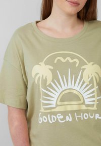 Olivgrünes Baumwoll-T-Shirt mit einem weißen und beigen Grafikdesign eines Sonnenmotivs, Wellen und Palmen sowie dem Text "Goldene Stunde."