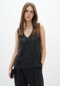 Top nero senza maniche con scollo a V, decorato con paillettes in motivi verticali, texture liscia, abbinato a pantaloni neri larghi.