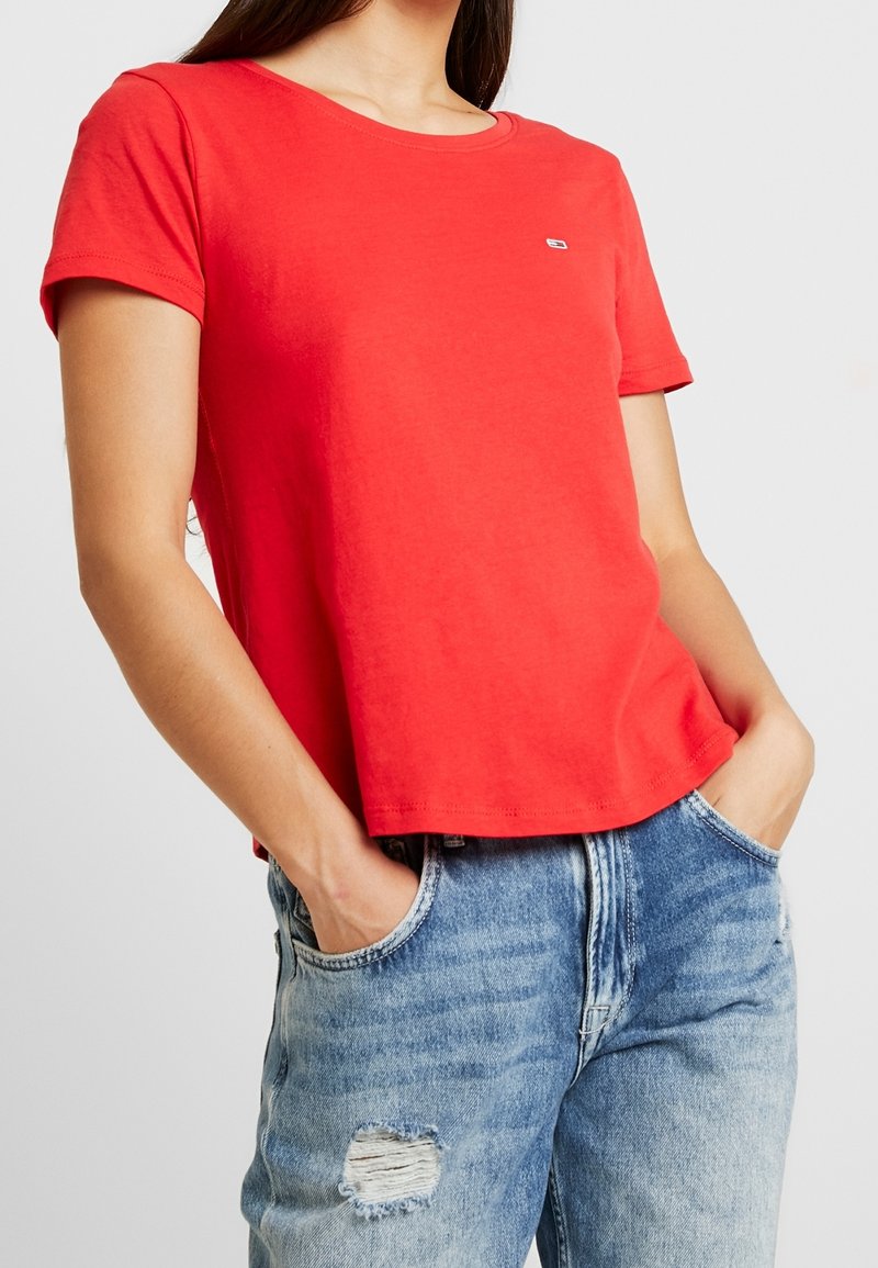 Röd bomullströja med rund nacke och korta ärmar. Har en liten logotyp på bröstet och är kombinerad med slitna blå denimjeans.