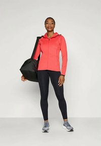 Kvinna i korallfärgad zip-up jacka, svarta leggings och grå sneakers står och håller en stor svart gymväska över axeln.