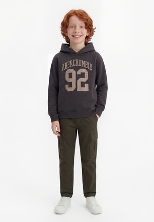 Χαμογελαστό αγόρι με κόκκινα μαλλιά φοράει μαύρο hoodie Abercrombie, χακί cargo παντελόνι και λευκά αθλητικά παπούτσια, στέκεται με το ένα χέρι στην τσέπη.