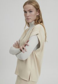 Top beige senza maniche con scollatura a V e spacchi laterali. Indossato sopra una camicia a maniche lunghe di colore chiaro. Texture liscia, vestibilità comoda.