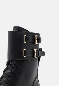 Bottine noire en cuir lisse avec une lanière à double boucle, une garniture en métal doré et un design à lacets sur le devant.