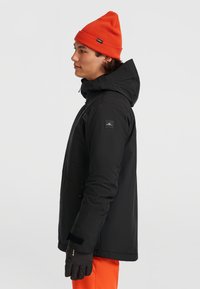 Zwarte waterdichte jas met een capuchon, zijzakken met rits en een logopatch. Gepaarde met een oranje muts en zwarte handschoenen.