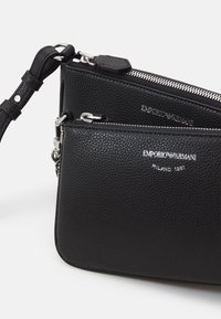 Černá texturovaná kožená crossbody taška se stříbrnými kovovými prvky, zipovým uzávěrem a vyraženým logem. Obsahuje nastavitelný popruh a kompaktní design.