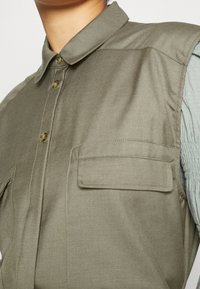 Zbliżenie na osobę noszącą oliwkowo-zieloną koszulę z guzikami, z kołnierzykiem i kieszonką klapą na piersi, na tle jasnoniebieskiego, teksturowanego rękawa.
