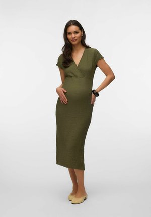 Femme enceinte en robe midi texturée vert olive à manches courtes, portant des ballerines beiges et un bracelet noir, posant sur fond blanc.