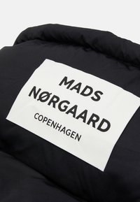 Czarny puchowy kurtka z białą metką, na której widnieje "MADS NØRGAARD COPENHAGEN" napisane grubymi czarnymi literami. Gładka tekstura, prostokątny kształt.