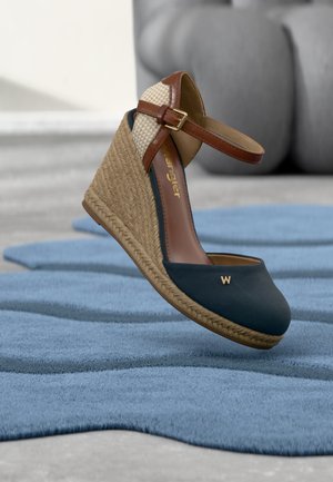 Dámská klínová bota s námořnicky modrým látkovým špičkou, podpatkem z tkaného juty, opásáním kotníku v barvě tan s přezkou a malým zlatým logem "W" na modrém texturovaném povrchu.