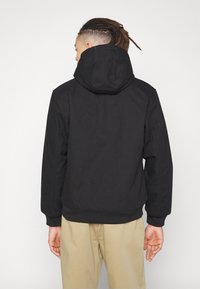 Black jacket with a hood, featuring a smooth, matte finish, ribbed cuffs, and a fitted design. Waist-length with a clean, minimal silhouette.