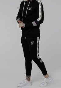 Felpa nera con zip, cordino bianco e logo. Pantaloni jogger neri abbinati con accenti logo laterali. Sneakers bianche completano l'outfit.