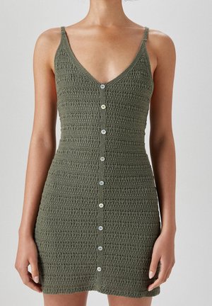 Vrouw draagt een olijfgroene mouwloze gehaakte minijurk met V-hals en een rij knopen aan de voorkant, gefotografeerd van schouder tot halverwege de dij.