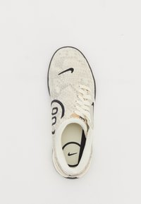 Zapatilla de color claro con una superficie texturizada, logo de Nike en negro, diseño con cordones y un interior acolchado. Presenta un acento circular cerca de los cordones.