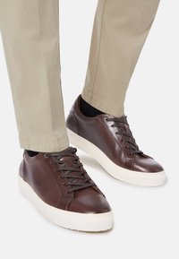 Braune Ledersneaker mit dunklen Schnürsenkeln, einer weißen Gummisohle und einer glatten Oberfläche. Getragen mit hellbeigen, taillierten Hosen.