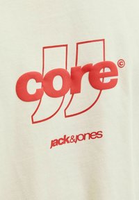 T-shirt en coton crème avec un texte rouge vif indiquant « core » et « jack & jones » avec un design distinctif de guillemets. Texture lisse.
