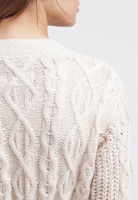 Detailní záběr na osobu v krémovém svetr s copánkovým vzorem, zobrazuje záda a část krku a brady.