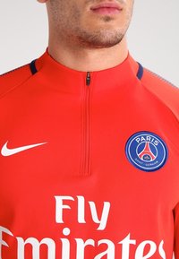 Chemise de sport Nike rouge avec une encolure zippée, arborant le logo « Fly Emirates » en blanc et l'écusson du Paris Saint-Germain en bleu.